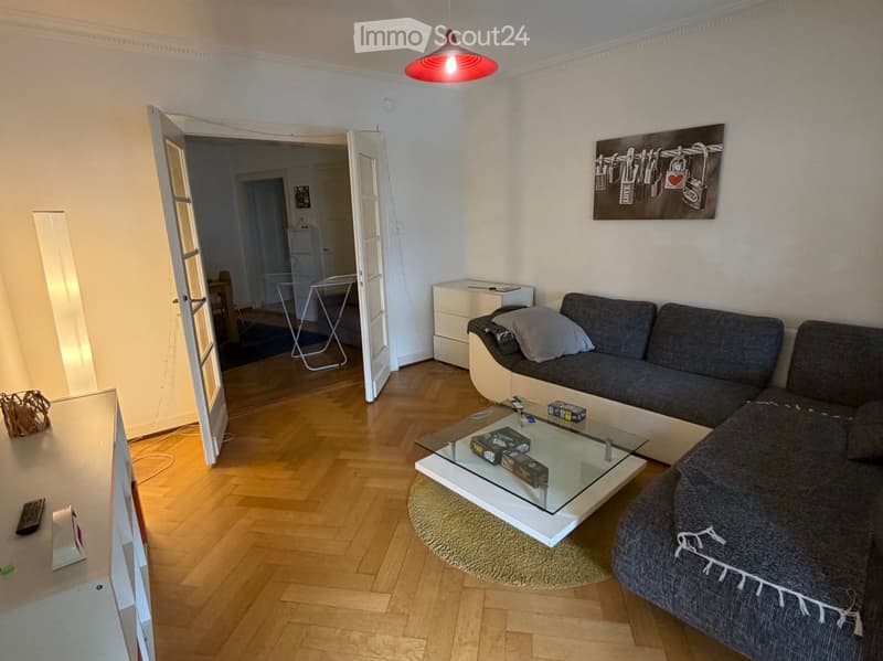 Modern living room with wooden flooring, grey sectional sofa, glass coffee table, and a large screen on the left side.