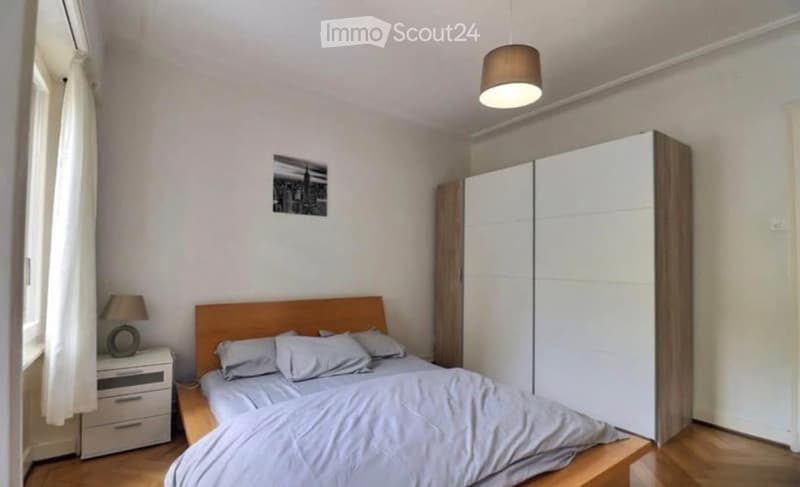 Modern bedroom with a wooden bed, nightstand, lamp, large wardrobe, and a cityscape picture on the wall.