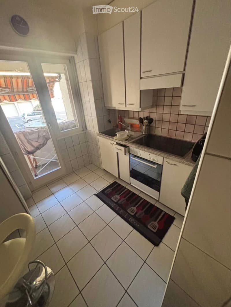 Compact kitchen with white cabinets, tiled walls, and a small window. Includes an oven, sink, and various kitchen utensils on the counter.