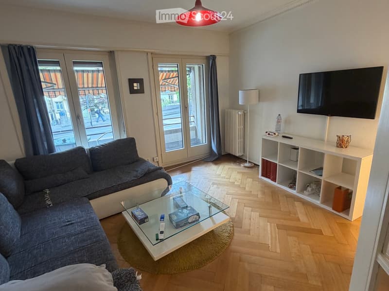 Modern living room with a grey sectional sofa, glass coffee table, white shelving unit, flat-screen TV, and a floor lamp. The room features large windows and a wooden floor.