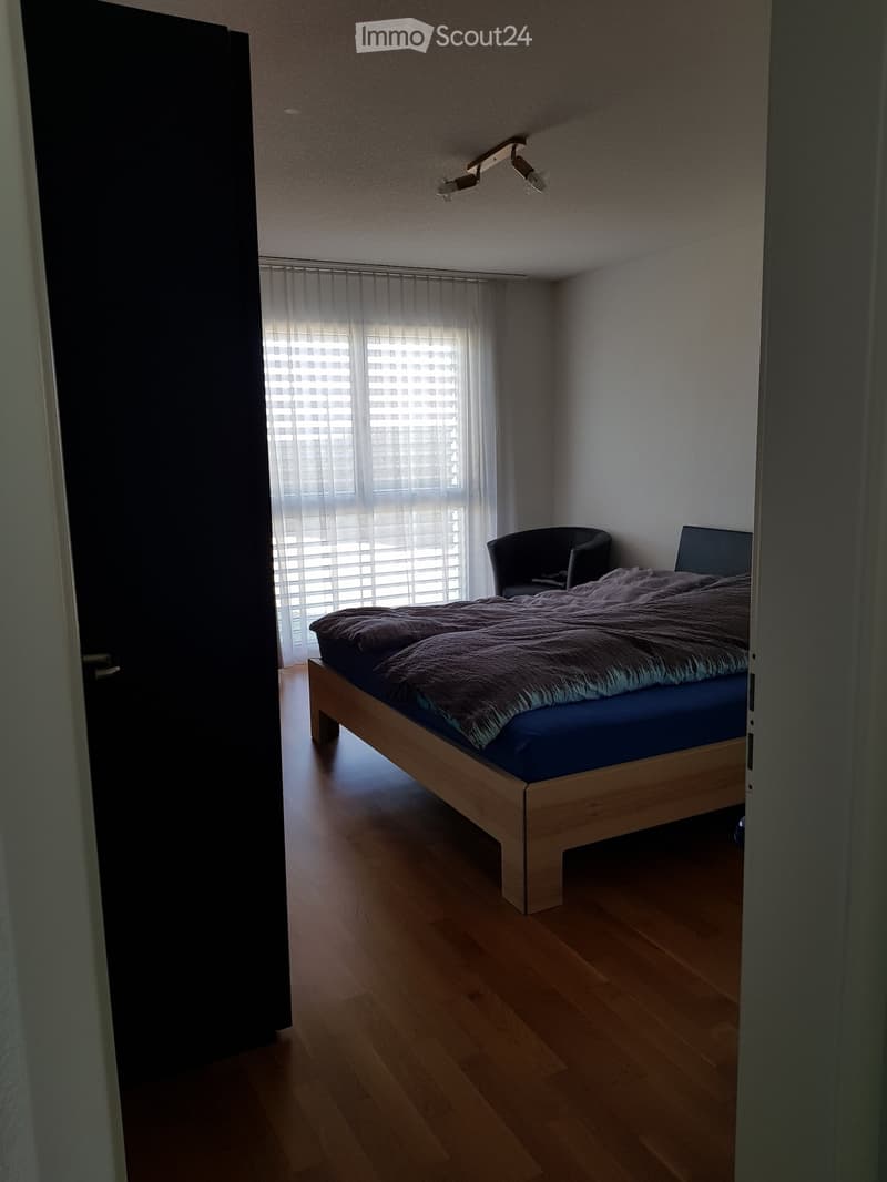 Minimalist bedroom with wooden floor, large window with blinds, bed with dark bedding, and a single chair.