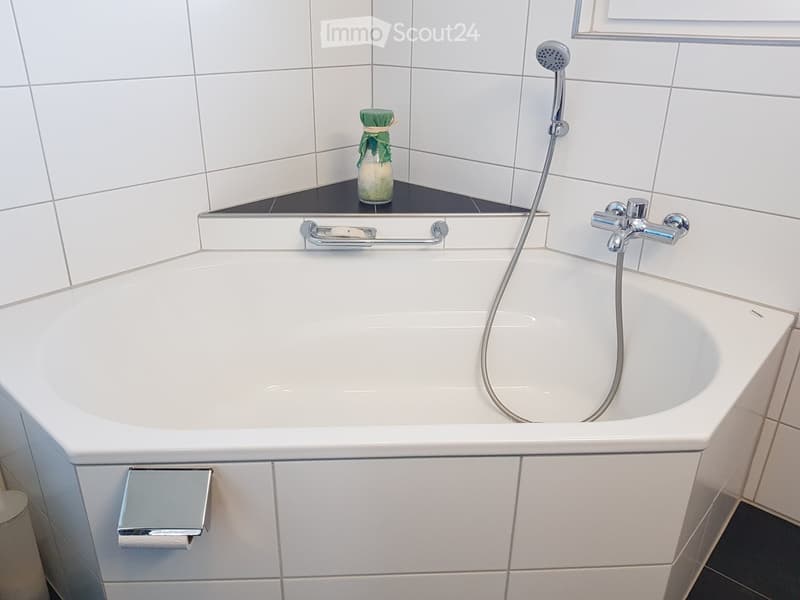 Modern bathroom with a white bathtub, chrome fixtures, a handheld showerhead, a mirror, and a small decorative bottle on the edge of the tub.