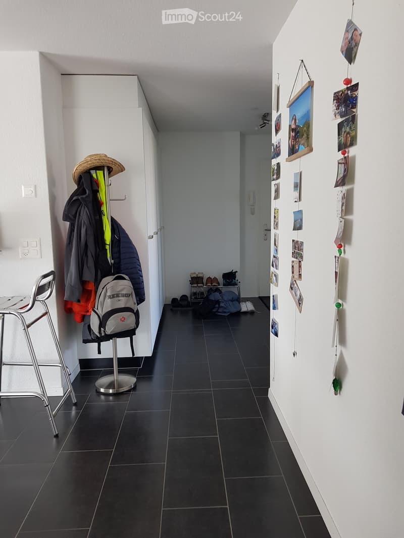 A narrow hallway with dark tiled flooring, white walls, and a coat rack holding jackets and a backpack. A small shelf with shoes and bags is visible at the end, and a string of photos and decorations hangs on the right wall.