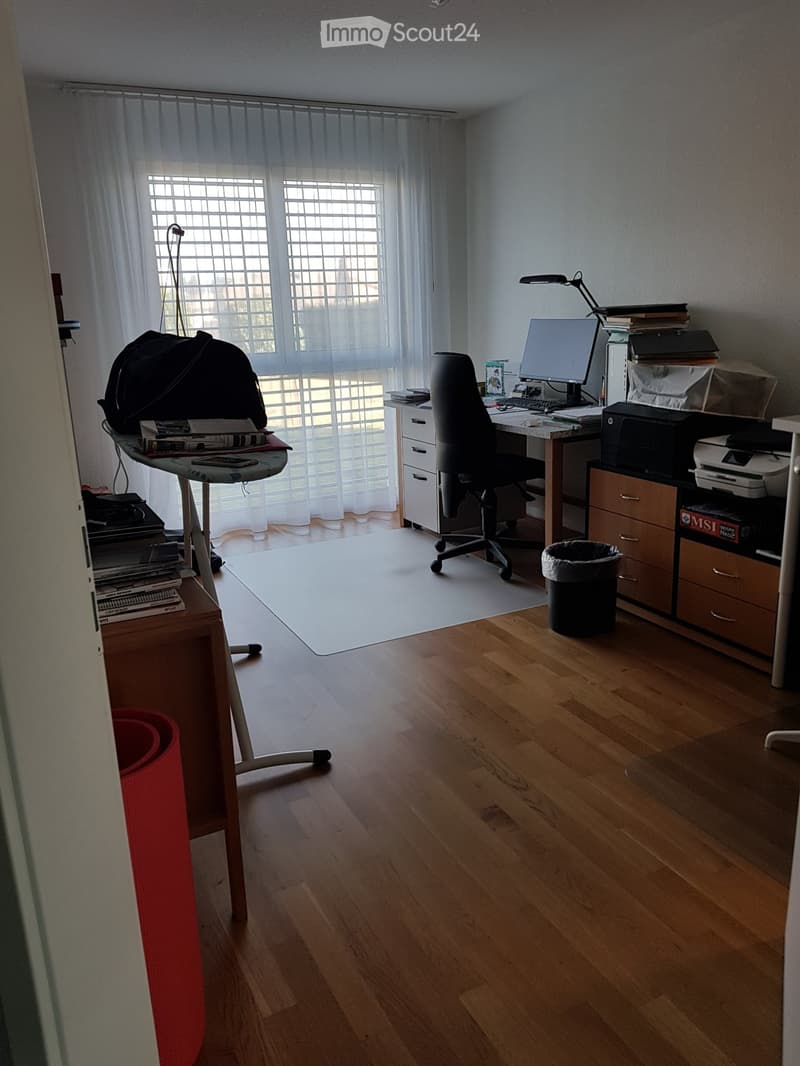 Well-lit office space with wooden flooring, large window with white curtains, desk with computer, chair, printer, and various office supplies.