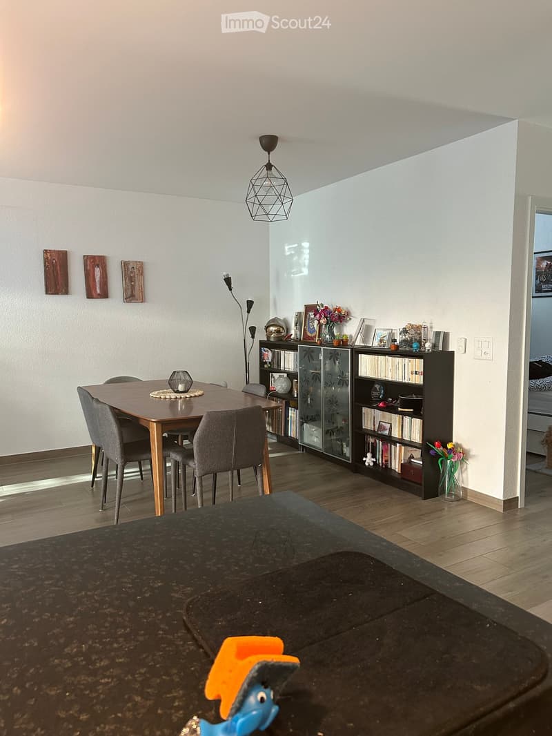 Modern dining area with a wooden table, grey chairs, bookshelf, and decorative items. The room features a pendant light, floor lamp, and artwork on the walls.