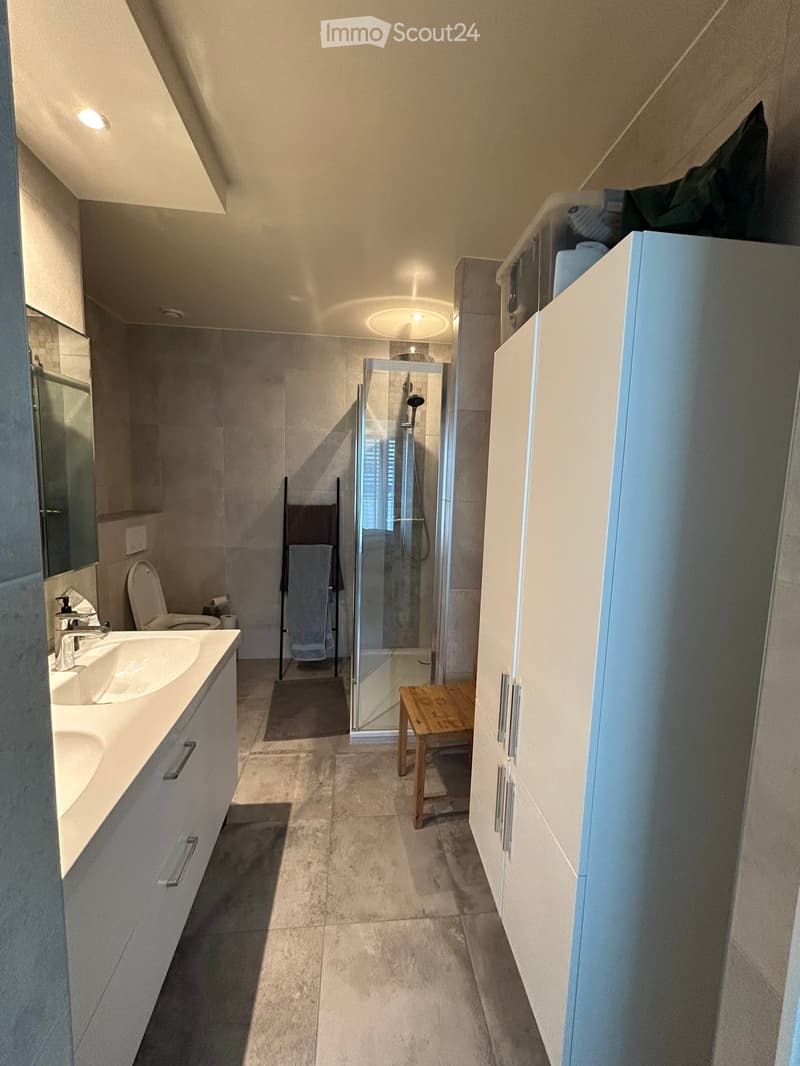 Modern bathroom with double sink, toilet, glass-enclosed shower, wooden stool, and towel rack with towels.