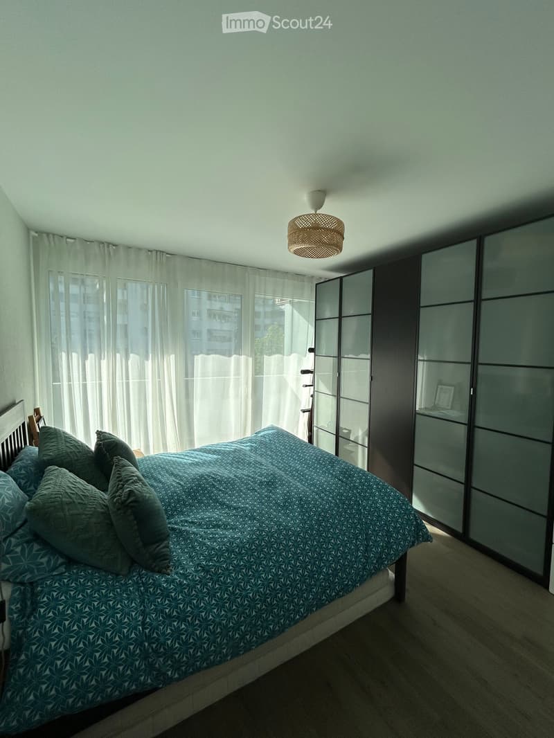 A modern bedroom with a large bed covered in a blue patterned duvet and matching pillows, positioned near a large window with white curtains. A sleek, dark-colored wardrobe with mirrored doors stands against the wall, and a woven ceiling light fixture han