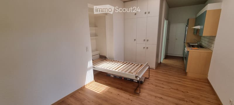 Empty room with wooden flooring, white walls, built-in wardrobes, and an adjacent kitchen area with cabinets and appliances.