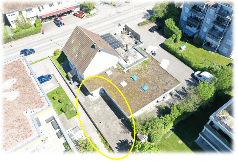 Aerial view of a multi-story residential building with a brown tiled roof, surrounded by greenery, parked cars, and neighboring structures.