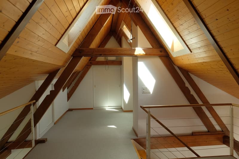 Spacious, well-lit hallway with wooden beams and slanted ceilings, featuring a staircase with wooden steps and metal railings leading to an attic or loft area.
