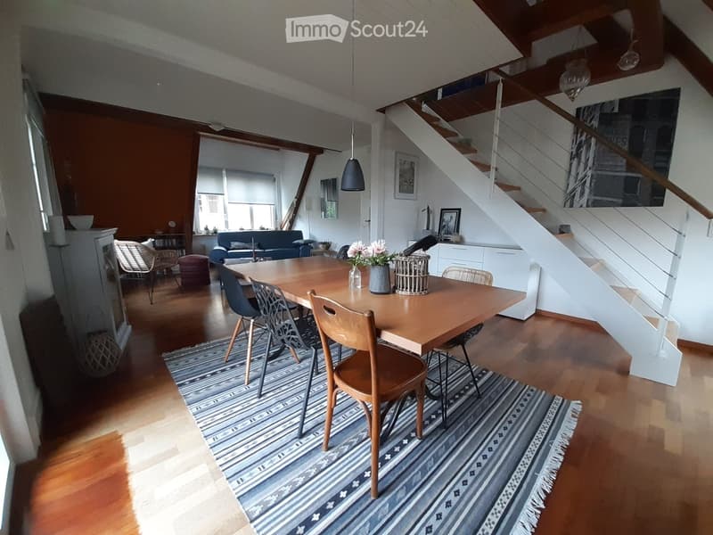 Bright living and dining area with wooden floors, a large dining table with chairs, a sofa, a staircase, and modern decor including a patterned rug and pendant lights.