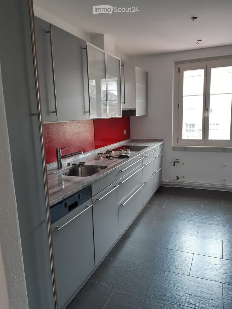Modern kitchen with grey cabinets, a red accent wall, a marble countertop, and a stainless steel sink.