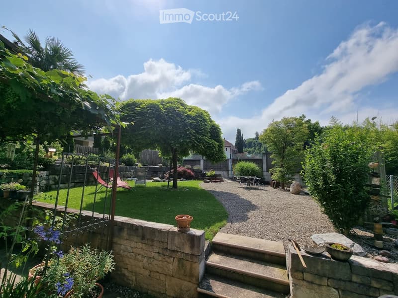 A well-maintained garden with a gravel pathway, a small lawn, potted plants, outdoor furniture, and a stone wall with steps leading up.