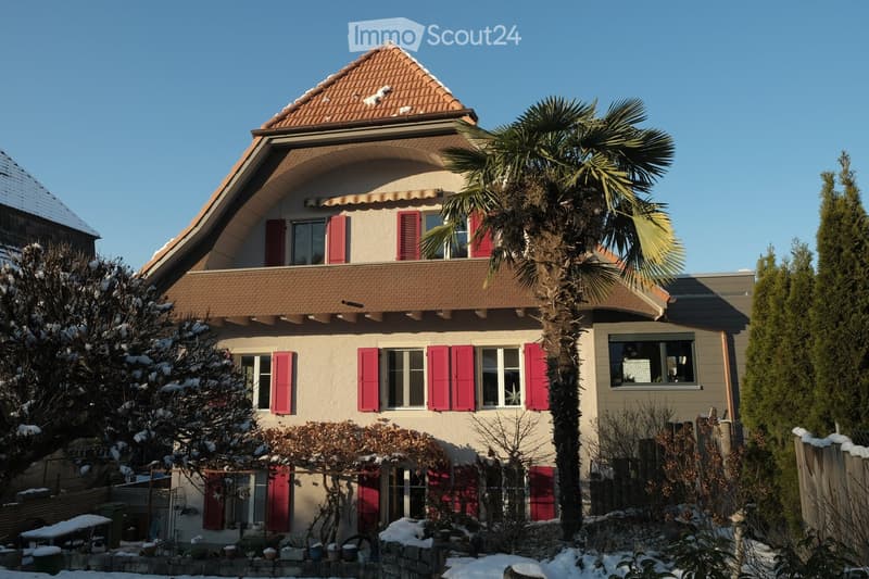 Two-story house with red shutters, beige walls, and a tiled roof. Snow is visible on the roof and surrounding trees. A palm tree is in the front yard.