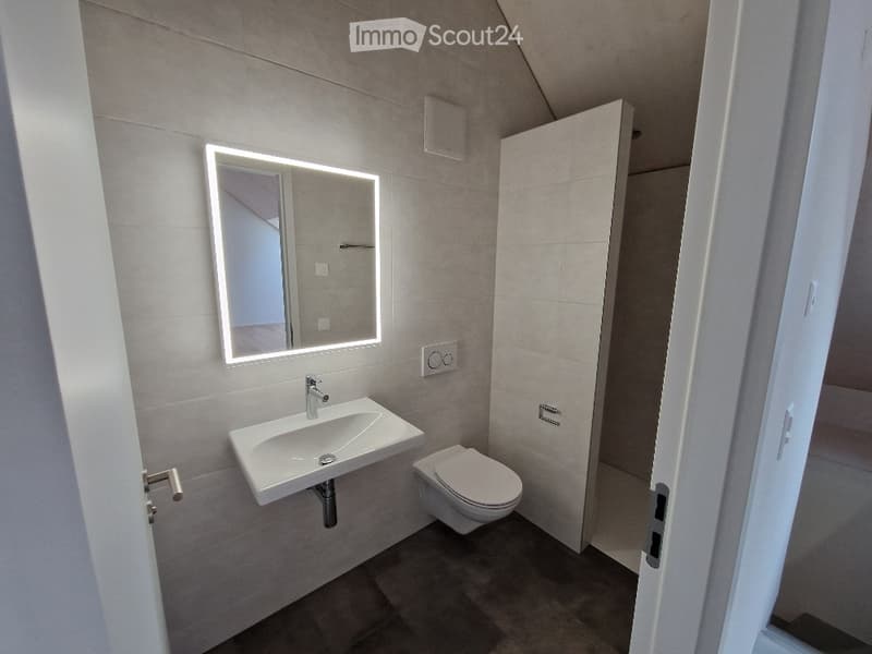 Modern bathroom with a white sink, chrome faucet, illuminated mirror, and a toilet. The walls are tiled in a neutral color, and the floor is dark.
