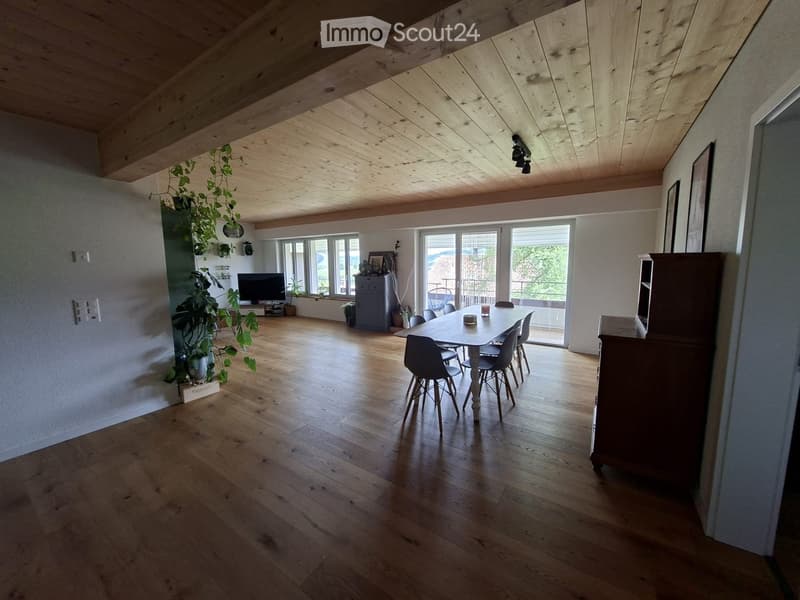 Open-concept dining and living area with wooden flooring, exposed wooden ceiling beams, large windows, and a sliding glass door leading to a balcony.