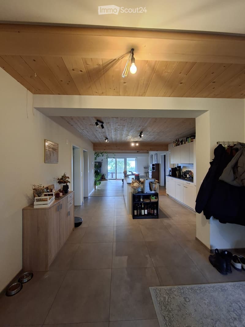 Bright hallway with wooden ceiling, tiled floor, and built-in storage units. Visible kitchen area with cabinets, appliances, and a small bar area with bottles. Shoes and jackets are placed near the entrance.