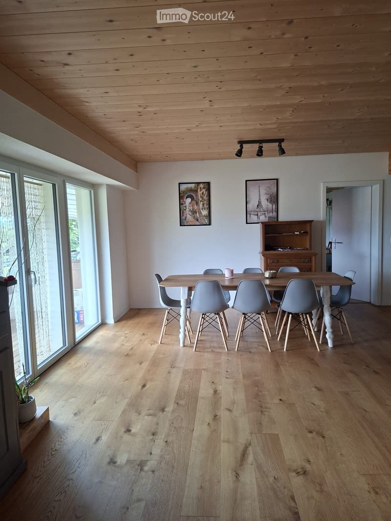 Bright dining area with wooden floor, white walls, and large windows. Features a dining table with six chairs, a cabinet, and two picture frames on the wall.