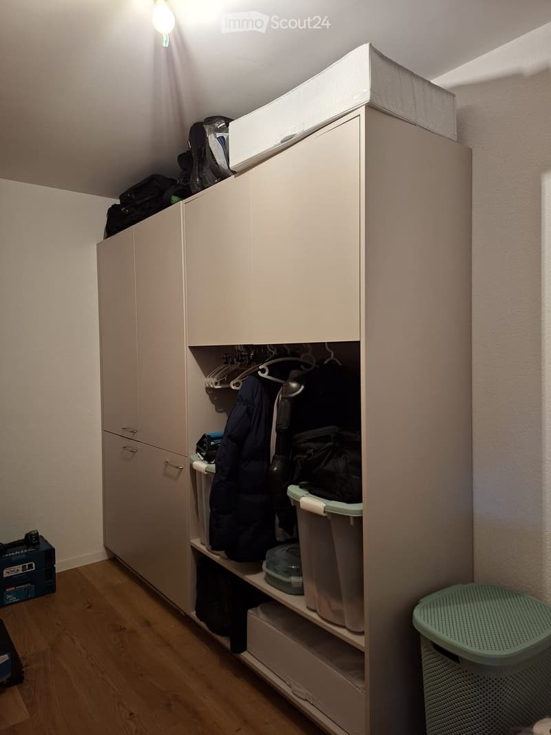 A small, well-lit closet with built-in white wardrobes and shelves holding various items, including bags, boxes, and a stool.