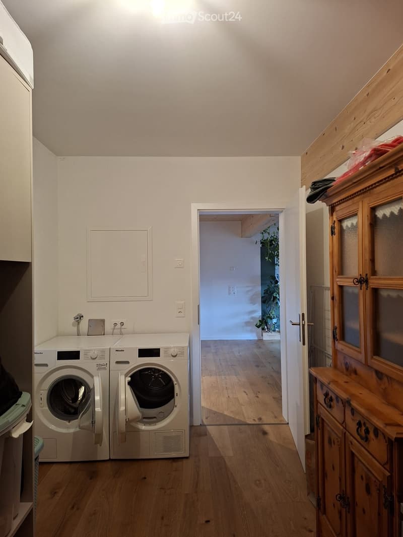 Hallway with wooden flooring, white walls, and a doorway leading to another room. Visible are a washing machine and dryer unit on the left side, and a wooden cabinet with drawers on the right side.