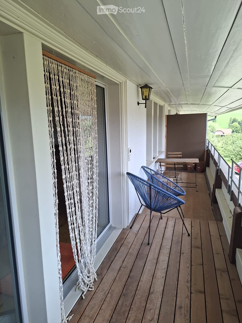 Wooden balcony with two blue chairs, a small table, and a hanging macrame curtain.