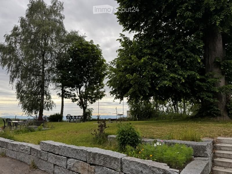 A serene garden area with a stone wall, grassy lawn, trees, and a few benches. The setting includes a view of the water in the background.