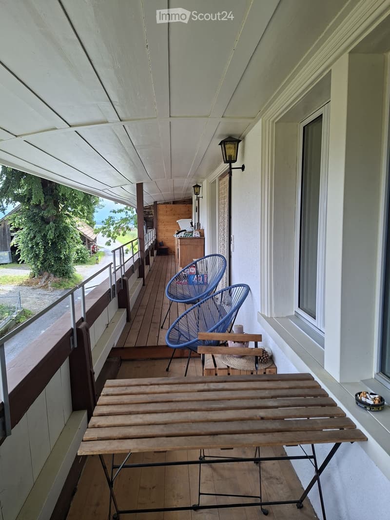 Balcony with wooden floor, metal-framed table, two blue wire chairs, small wooden side table, and outdoor lighting.