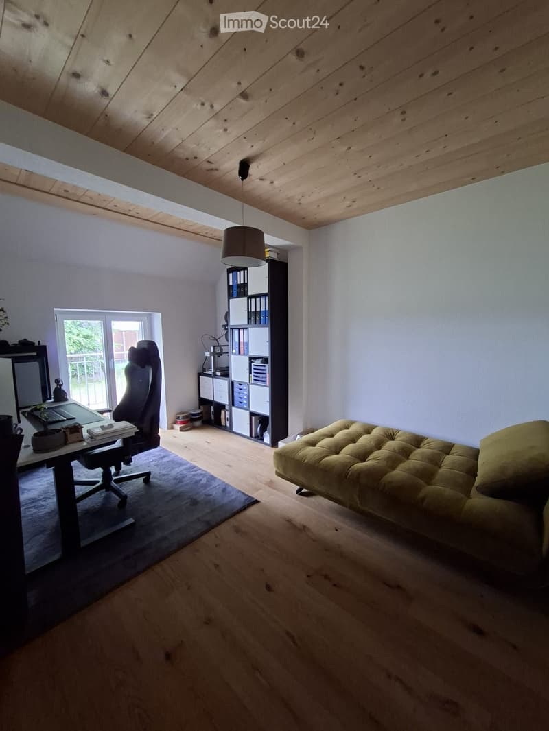 Bright, open room with wooden floors, a large window, a sofa, a chair, a table with a laptop, and a shelf with books and storage boxes.