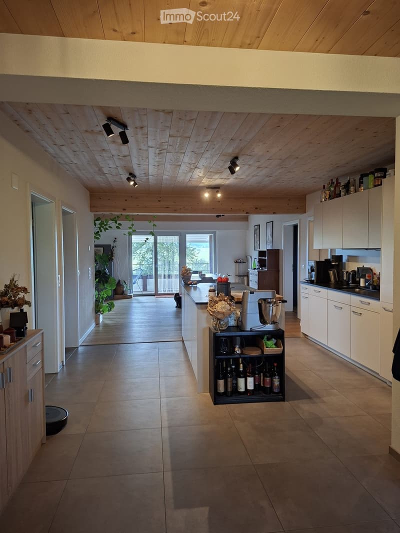 Modern kitchen with white cabinets, stainless steel appliances, and a wine rack. The space features a large island with a countertop, a coffee maker, and a blender. There is a cat visible in the background, along with potted plants and framed pictures on