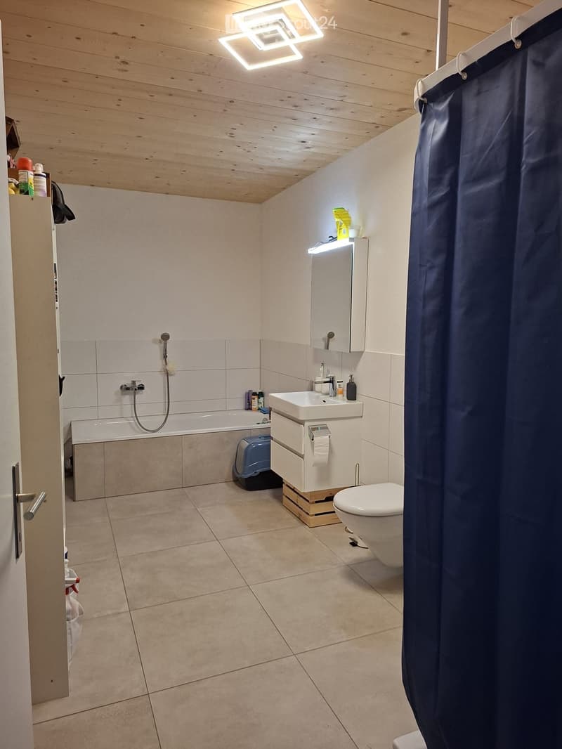 Modern bathroom with a bathtub, sink, toilet, and mirror. Features a shower curtain, tiled walls, and a wooden ceiling with integrated lighting.