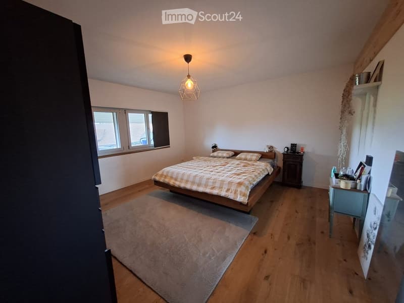 Cozy bedroom with wooden flooring, a large bed with a checkered duvet, a nightstand, a small table with various items, and a hanging geometric lamp.