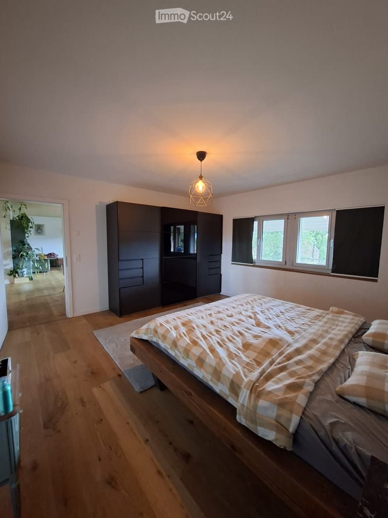 Modern bedroom with wooden flooring, a large bed with a checkered duvet, a tall dark wardrobe, and a hanging pendant light fixture.