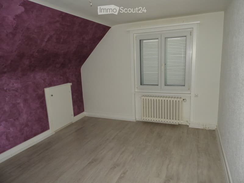 Empty room with wooden flooring, white walls, and a purple accent wall. Features a window with white shutters and a radiator below it.