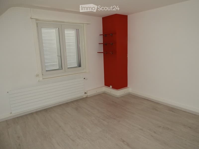 Empty room with light wooden flooring, white walls, a window with white shutters, a white radiator, and a red accent wall with built-in shelves.