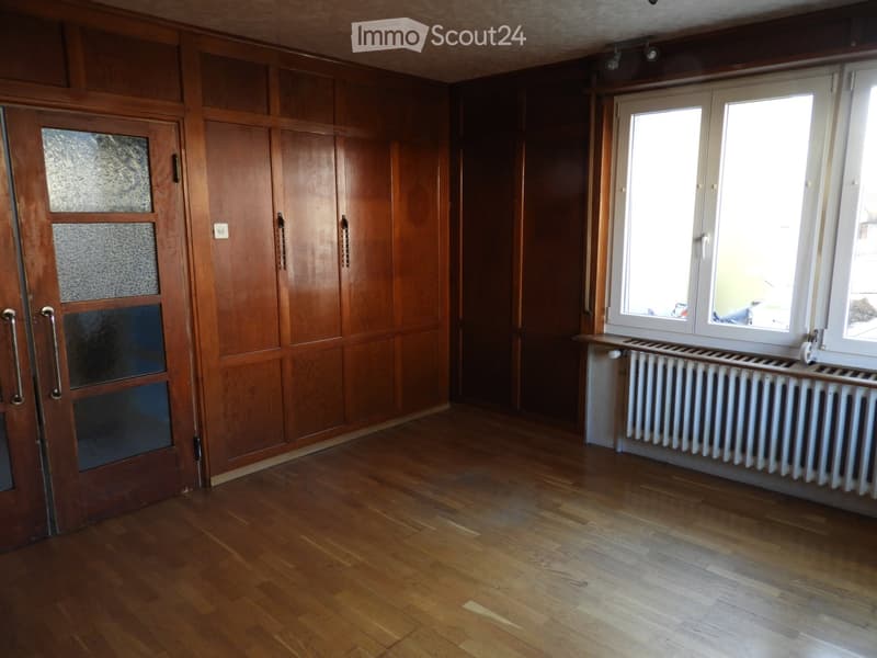 Empty room with wooden walls, wooden floor, large windows, and a radiator under the windows.