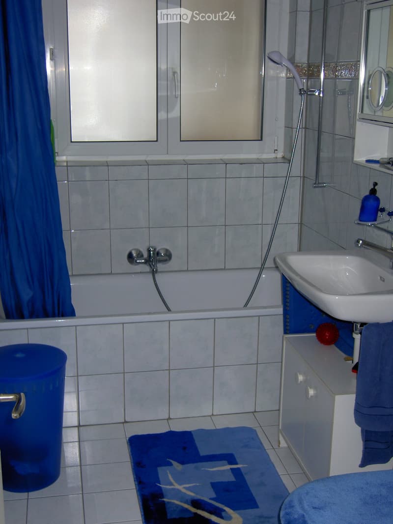 Bathroom with a bathtub, sink, mirror, and towel. Features tiled walls and floor, a window with a blue curtain, and a small cabinet next to the sink.
