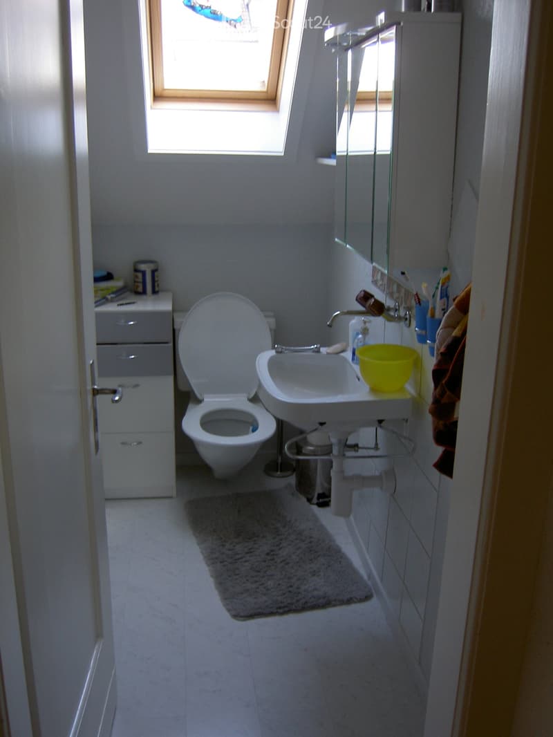 A small, clean bathroom with a toilet, sink, mirror, and a small cabinet. The room has white tiled walls and floor, a grey rug, and a window letting in natural light.