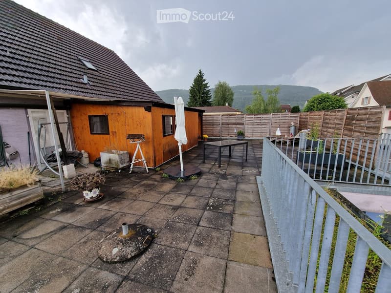 Outdoor patio with tiled floor, wooden fence, small table and chairs, umbrella, potted plants, and a small wooden shed.