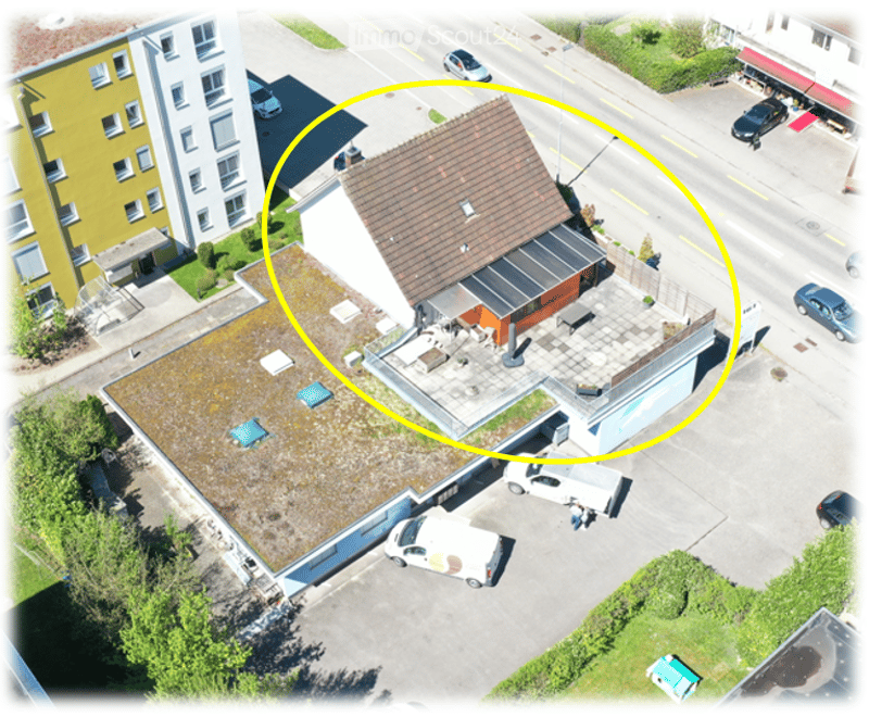 Aerial view of a multi-story building with a flat roof, surrounded by a parking lot and other structures. The building has a modern design with visible windows and a small garden area on the roof.
