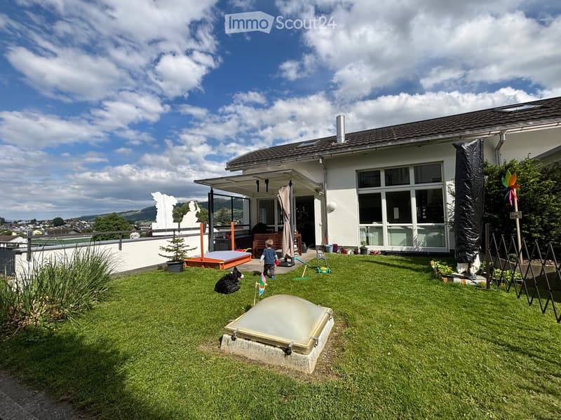 Lush green garden with a modern house in the background, featuring a patio area, a small wooden bench, a dog, a child, and a covered outdoor seating area.