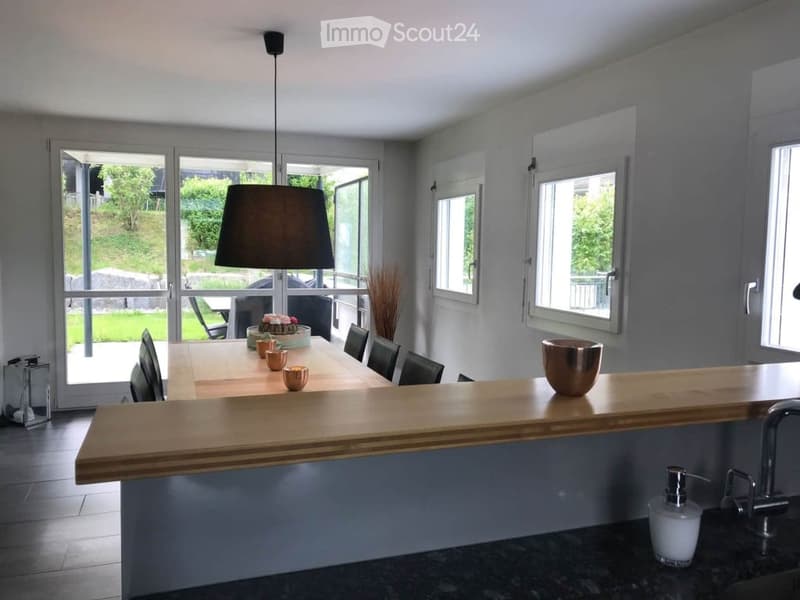 Modern dining area with a long wooden table, chairs, and a bar counter. The room features large windows with blinds, a pendant light, and decorative items on the table and counter.
