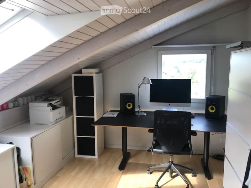 A modern attic office with wooden floors, slanted ceilings, and large windows. Features a black desk with a monitor, chair, lamp, and speakers, along with a white shelf and printer.
