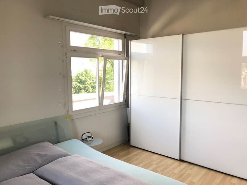 Modern bedroom with a light blue bed, grey bedding, a white wardrobe, and a small nightstand with a lamp and speaker.