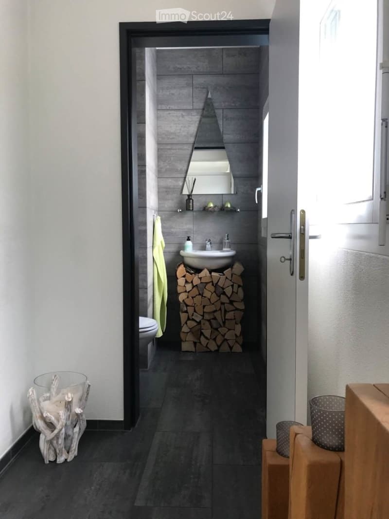 Modern bathroom with dark grey tiles, a sink on a wooden block, a mirror, a toilet, and a towel hanging on the wall. A wooden stool and a trash bin are visible in the foreground.