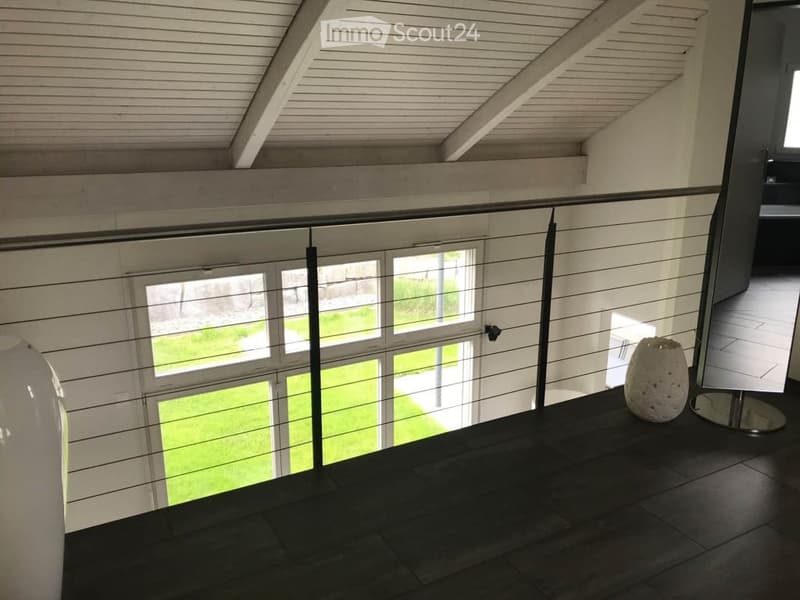 Modern hallway with a sleek glass railing, wooden ceiling beams, and a view of a green outdoor area through large windows.
