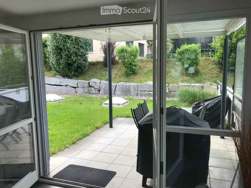 Open patio with tiled floor, covered roof, covered grill, chairs, and a view of a well-maintained garden with a stone wall and greenery.