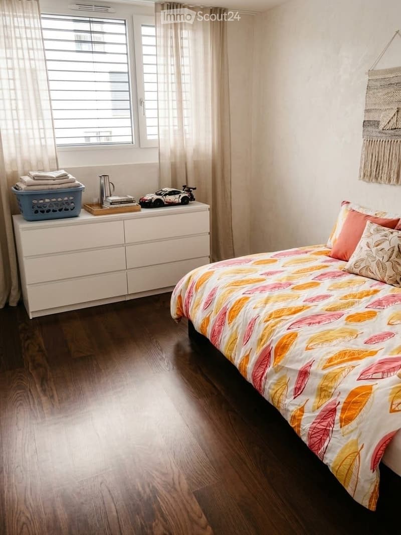 A modern bedroom with a large bed, colorful bedding, a nightstand with books and a toy car, a drawer unit, and a window with curtains.