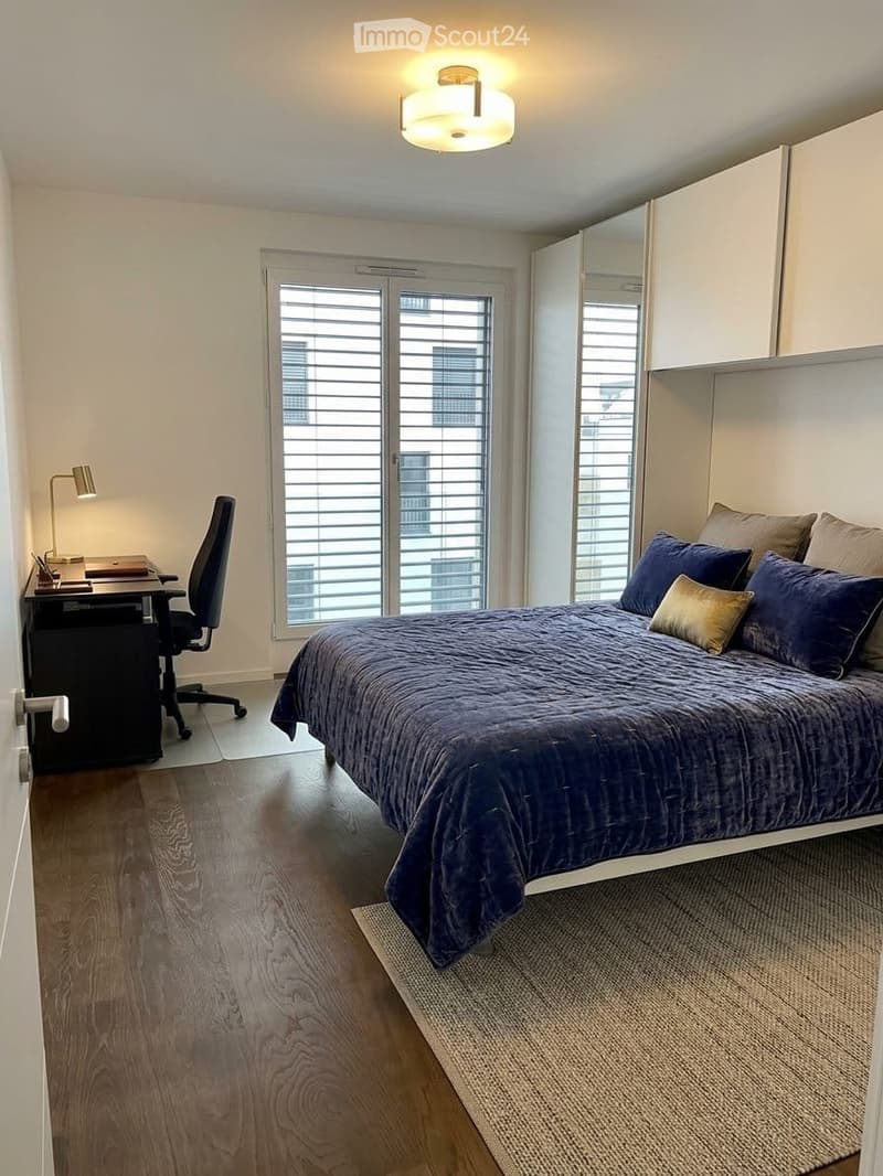 Modern bedroom with a large bed, desk, chair, and large windows with blinds. The room features a wooden floor, a ceiling light fixture, and a built-in wardrobe with a mirror.