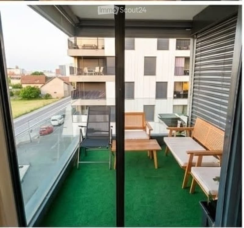 Balcony with green artificial turf flooring, wooden furniture including a bench, small table, and chairs, overlooking a street with parked cars and neighboring buildings.