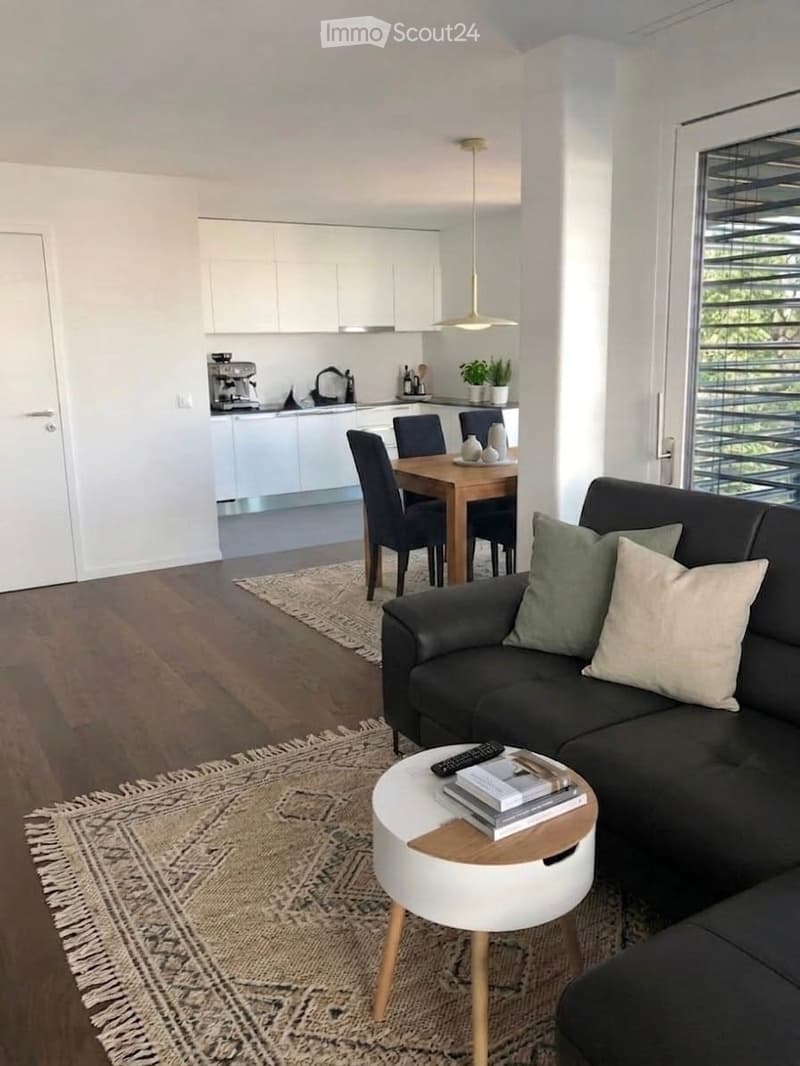 Modern open-plan living and dining area with a dark grey sofa, white coffee table, dining table with chairs, and a small kitchen area with white cabinets and a coffee machine.